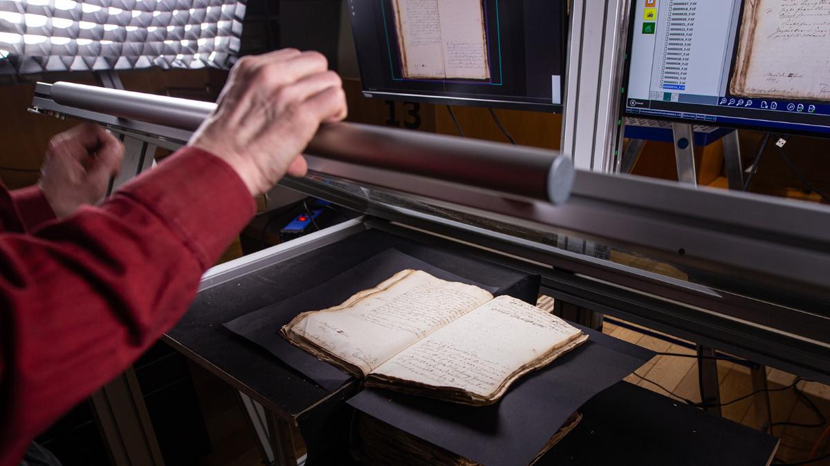 Kuvassa digitoidaan aineistoa: pöydällä on avonainen vanha aineisto, josta otetaan kuvaa lasin läpi