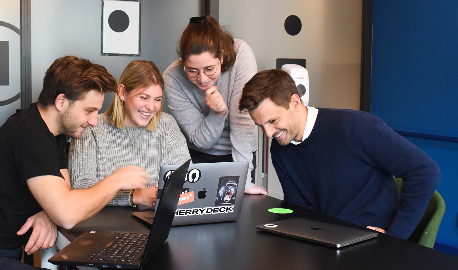 Kuvassa neljä nuorehkoa ihmistä katsomassa iloisena samaa kannettavaa tietokonetta.